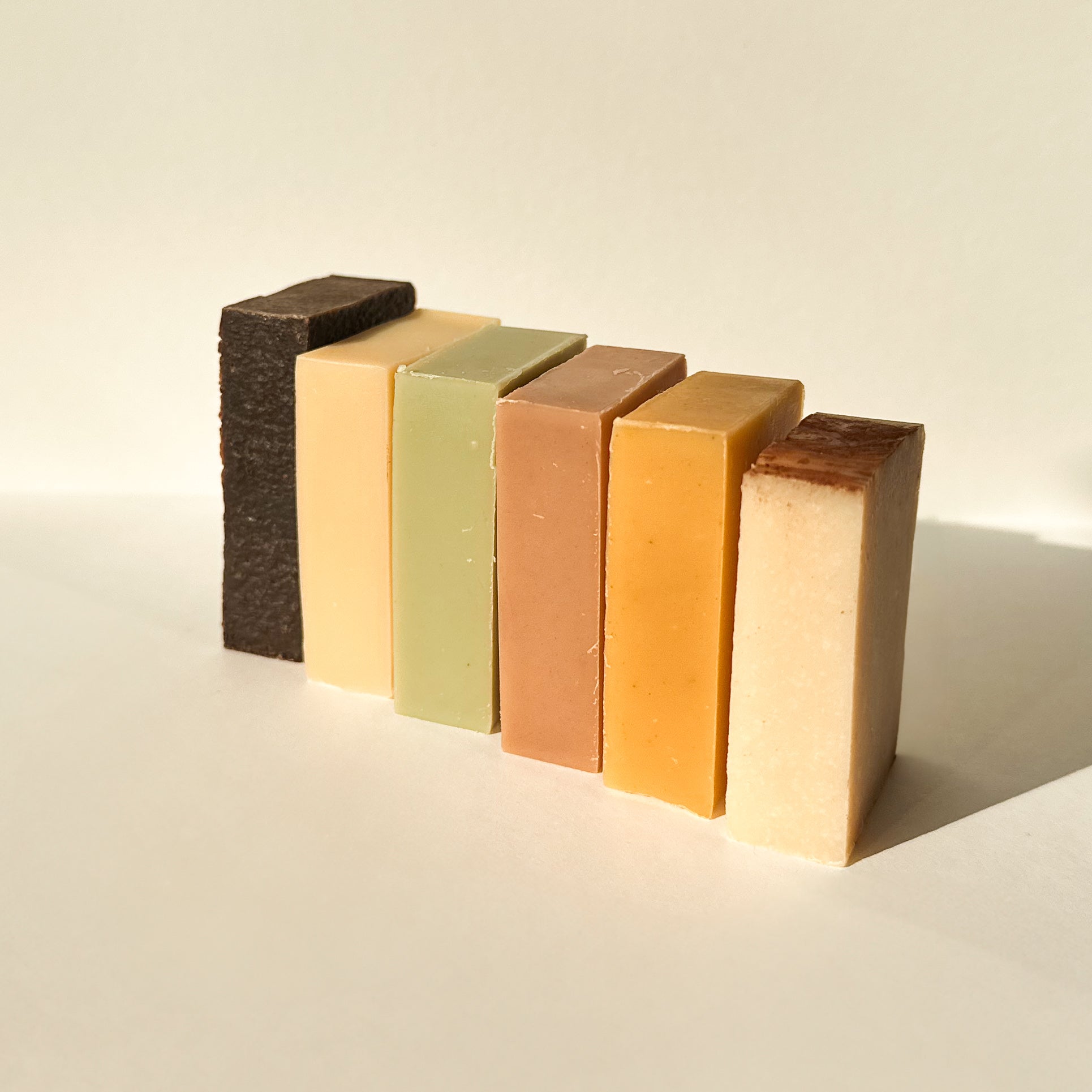 Six natural, handmade mini soap bars from NÜSKA, arranged in a row. Each soap bar has a distinct color, reflecting its natural ingredients, such as green, beige, orange, and brown. The soaps are simple and minimalist, showcasing a variety of scents, perfect for eco-conscious self-care routines.