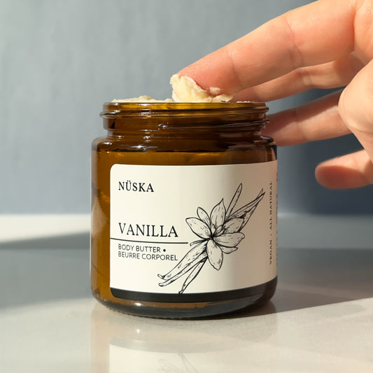 Hand picking up a small amount of Nüska Vanilla Body Butter from an amber jar showing its smooth, moisturizing texture.