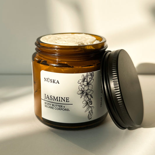 Nüska Jasmine Body Butter in an Open Amber Glass Jar with a Black Lid on the Side.