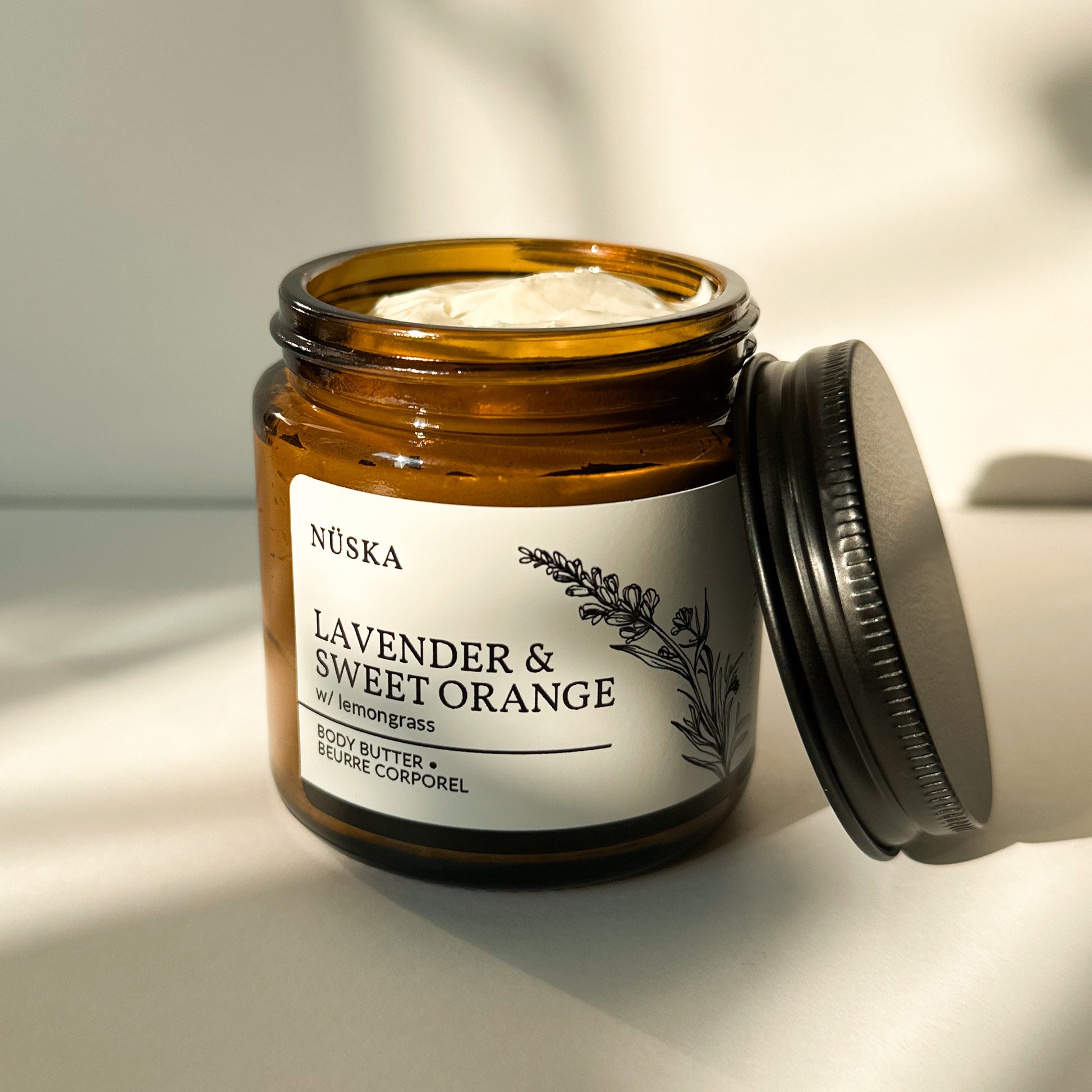 Nüska Lavender and Sweet Orange Body Butter in an Open Amber Glass Jar with a Black Lid on the Side.