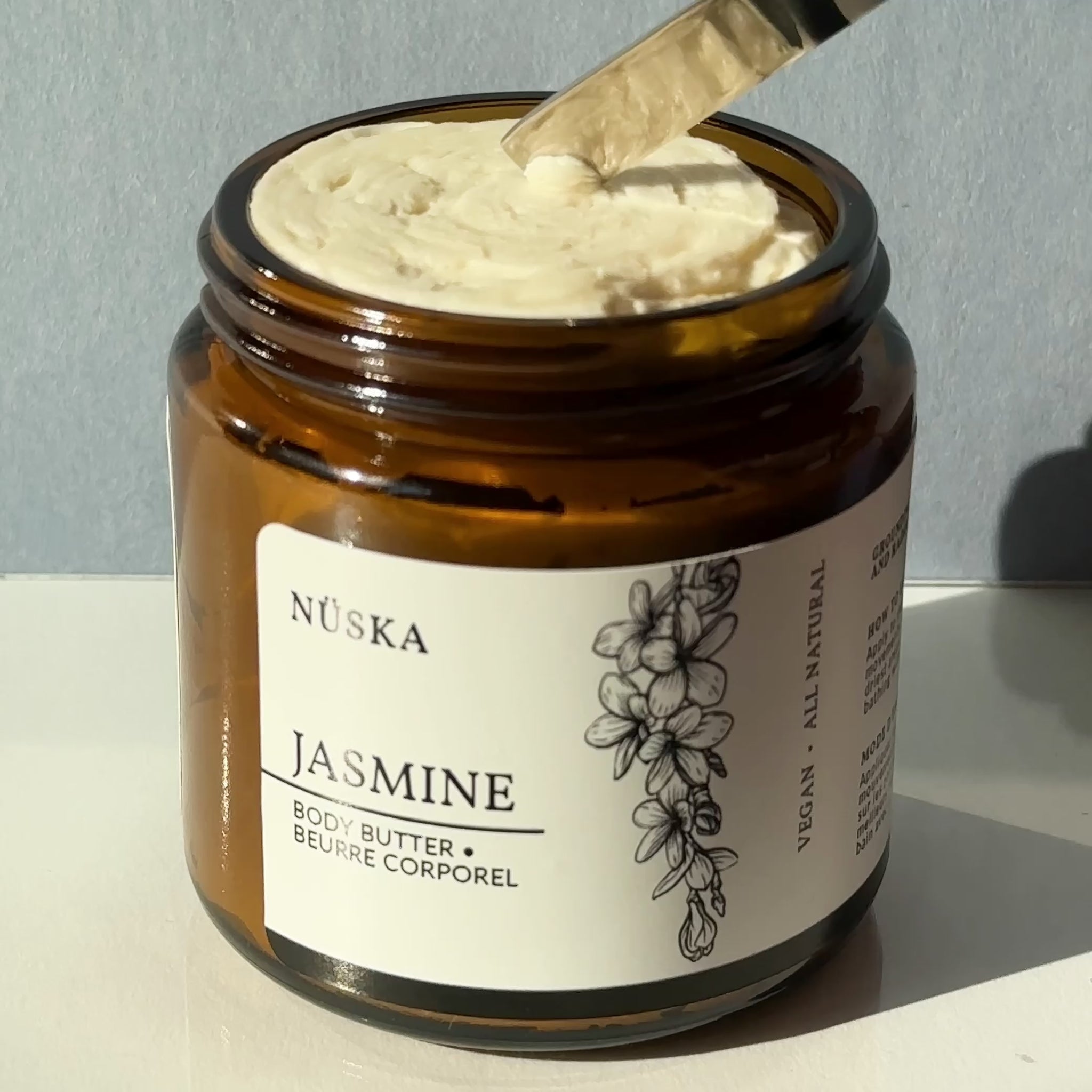 Nüska Jasmine Body Butter in an open amber glass jar, with a spatula scooping the creamy texture for a close-up view of the rich, nourishing butter.